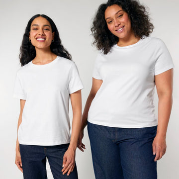 Two smiling women wearing white STTW172 Stella Muser The Iconic Womens T-Shirts made of soft organic cotton by Stanley/Stella and blue jeans stand side by side against a plain background.