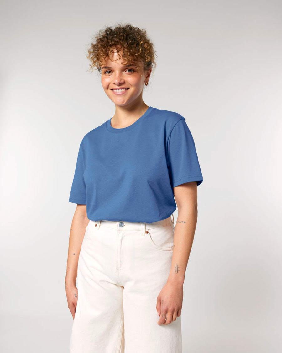 A person with curly hair smiles while wearing the STTU170 Stanley/Stella Crafter The Iconic Mid-Light Unisex T-Shirt in blue and white pants, standing against a plain background.