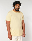 A man with short curly hair and a beard, wearing the iconic STTU170 Stanley/Stella Crafter The Iconic Mid-Light Unisex T-shirt in light yellow made of organic cotton and light-colored pants, stands against a plain background.