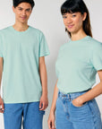 Two individuals, one male and one female, stand side by side wearing matching light green STTU170 Stanley/Stella Crafter The Iconic Mid-Light Unisex T-shirts made from organic cotton and blue jeans, both smiling at the camera.