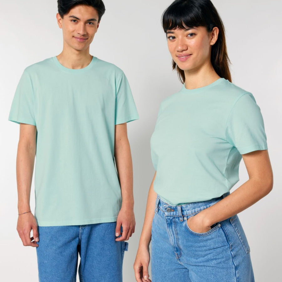 Two individuals, one male and one female, stand side by side wearing matching light green STTU170 Stanley/Stella Crafter The Iconic Mid-Light Unisex T-shirts made from organic cotton and blue jeans, both smiling at the camera.