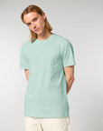 A person with long blonde hair wearing an iconic Stanley/Stella Crafter The Iconic Mid-Light Unisex T-shirt made of organic cotton in light green and off-white pants stands against a plain background.