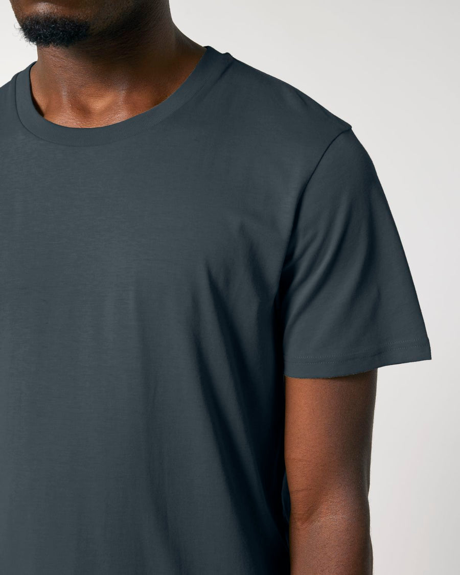 A person wearing the Stanley/Stella Crafter The Iconic Mid-Light Unisex T-shirt in plain dark grey, made from organic cotton, stands against a light grey background. The photo shows the upper body and part of the face.