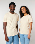 A man and a woman wearing iconic beige STTU170 Stanley/Stella Crafter The Iconic Mid-Light Unisex T-shirts made from organic cotton and blue jeans stand side by side, both smiling at the camera against a plain background.