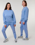 Two people wearing garment-dyed light blue MyNeedsAreSimple STSU085 Stanley Stella Matcher Vintage Unisex Medium Fit Crew Neck Sweatshirts pose against a plain white background. Both are standing, facing the camera, and smiling in their unisex sweatshirts made from organic cotton.