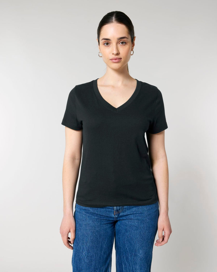 A person with dark hair tied back is wearing a plain black STTW176 Stella Isla Womens V Neck T-Shirt by Stanley/Stella, made of organic cotton, along with blue jeans, standing against a light gray background.