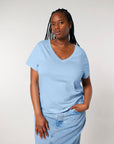 A woman with braided hair wears a light blue STTW176 Stella Isla Women's V-Neck T-Shirt by Stanley/Stella, made from organic cotton, along with light-wash denim jeans against a plain background.