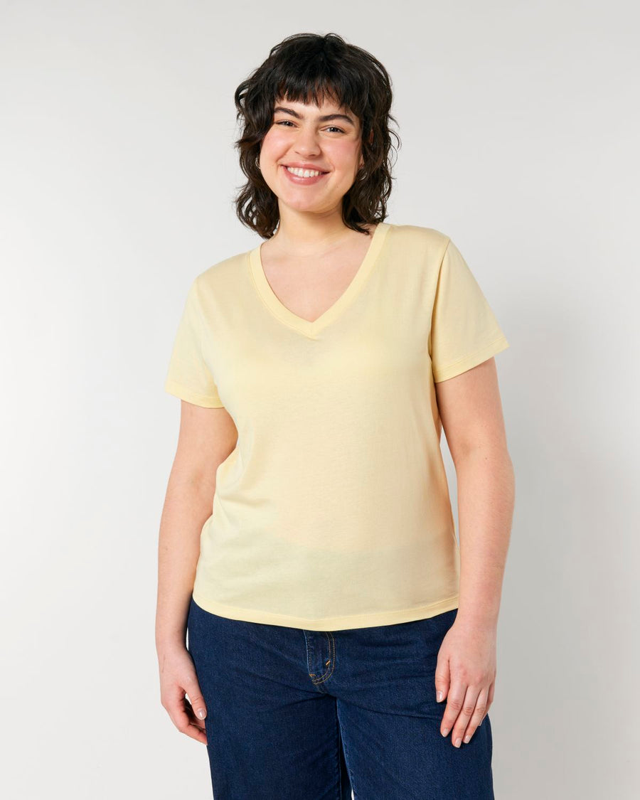 A person with short dark hair is smiling and wearing a light yellow STTW176 Stella Isla Women's V-Neck T-Shirt by Stanley/Stella, made of organic cotton, paired with dark blue jeans. They are standing against a plain white background.