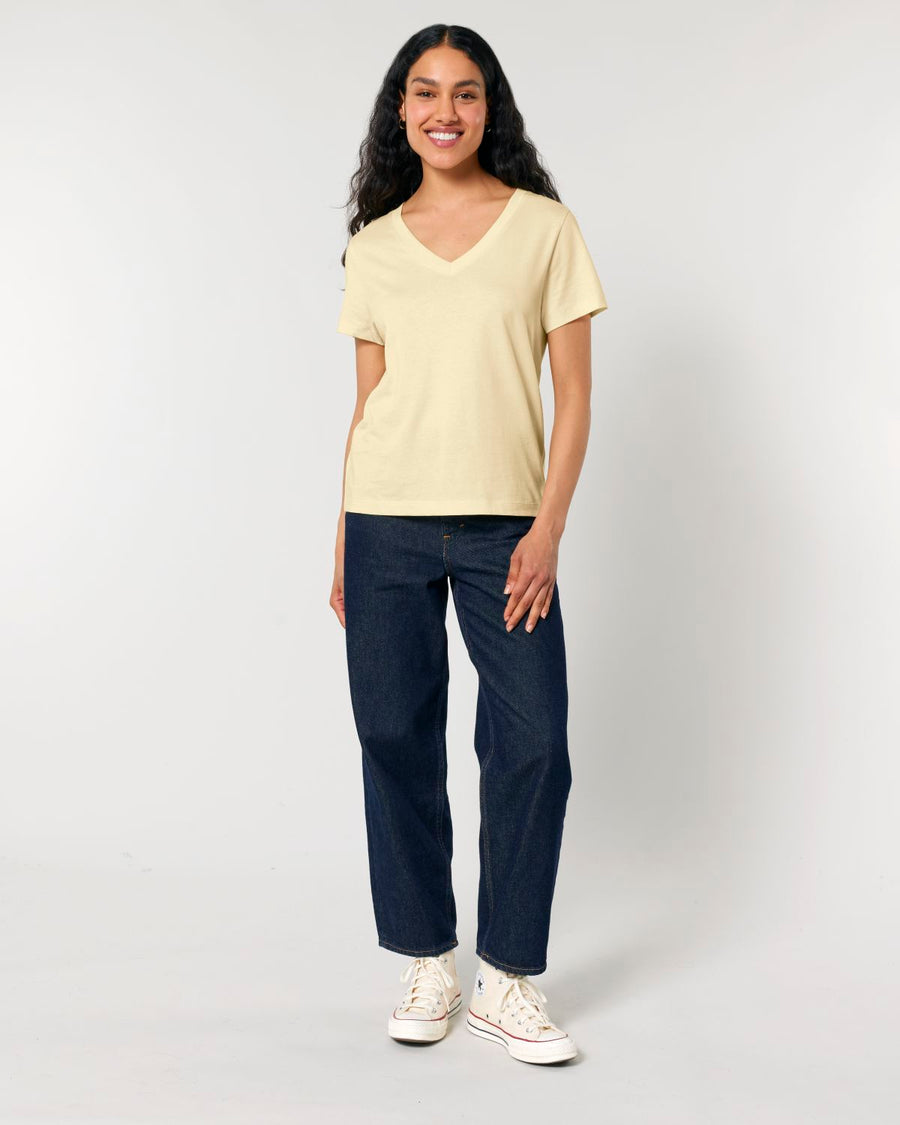 A woman with long, dark hair stands against a plain background, wearing a light yellow STTW176 Stella Isla Womens V Neck T-Shirt by Stanley/Stella made from organic cotton and dark blue jeans. Her white sneakers complement her relaxed stance as she smiles gently.