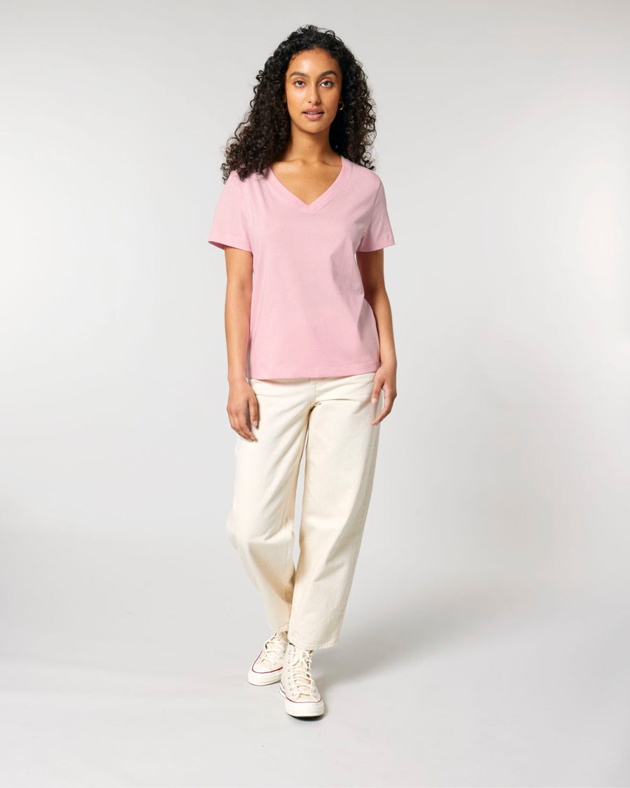 A person with long curly hair wears a pink Stanley/Stella STTW176 Stella Isla Women's V Neck T-Shirt made from organic cotton, cream-colored trousers, and white high-top sneakers, standing and looking forward in a studio with a plain background.