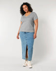 A person with curly hair, wearing a gray STTW176 Stella Isla Women's V Neck T-Shirt by Stanley/Stella made from organic cotton, a long denim skirt with a front slit, and white sneakers is standing against a plain background.