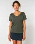 A person stands against a plain background, wearing an STTW176 Stella Isla Women's V-Neck T-Shirt by Stanley/Stella in organic cotton green and a dark denim skirt, with a neutral expression and hands at their sides.