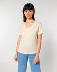 A person with shoulder-length dark hair wearing a cream-colored STTW176 Stella Isla Womens V Neck T-Shirt by Stanley/Stella, made from soft, single jersey organic cotton, and blue jeans stands against a plain white background.