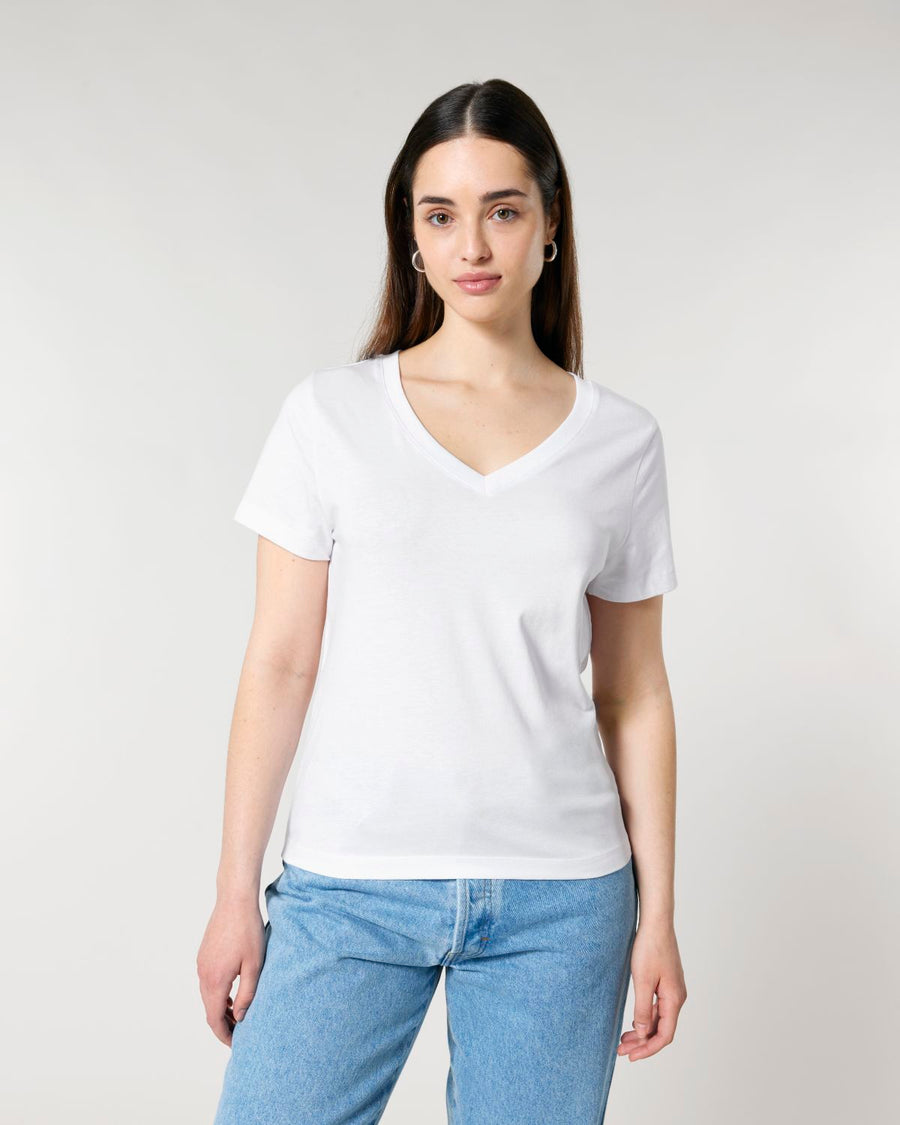 A woman wearing a white STTW176 Stella Isla Womens V Neck T-Shirt from Stanley/Stella and blue jeans stands against a plain, light-colored background. The SINGLE JERSEY fabric of the shirt offers a soft, comfortable fit.