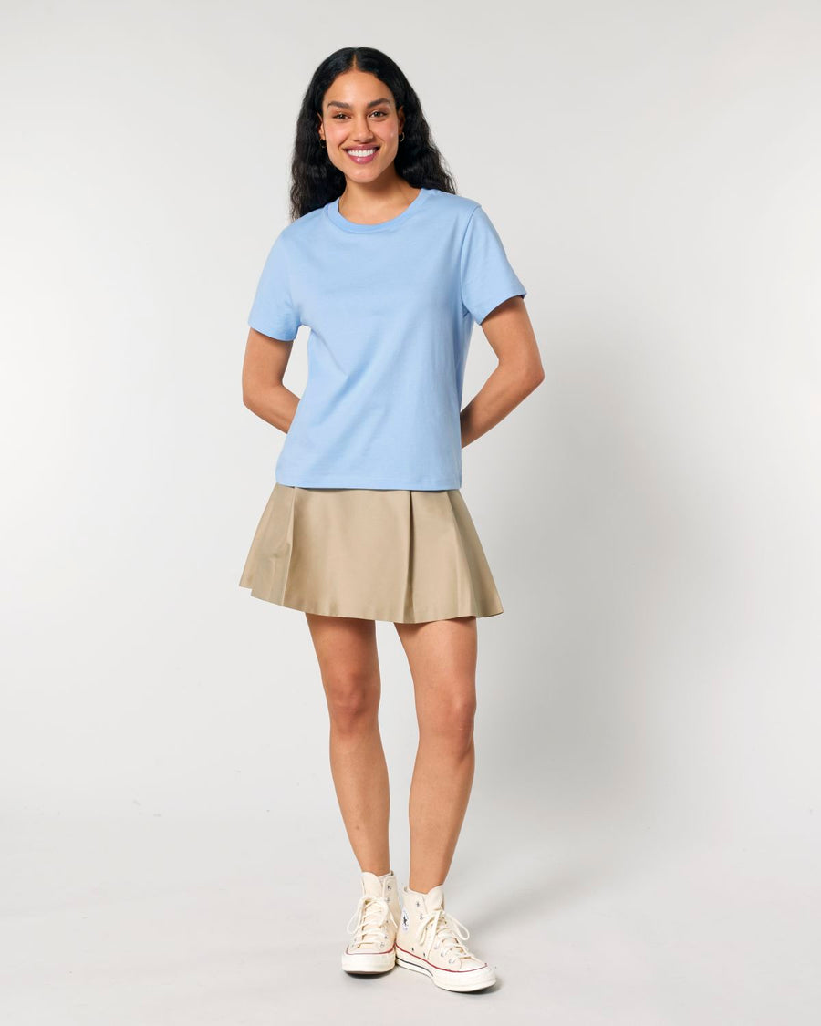 A person with long dark hair is standing against a plain background, wearing a Stanley/Stella STTW172 Stella Muser The Iconic Womens T-Shirt, a beige skirt, and white sneakers.