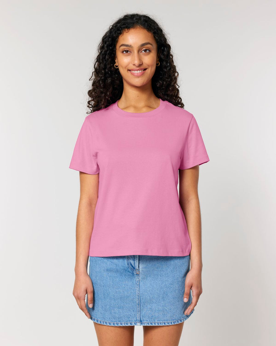 A person with long curly hair is wearing a pink STTW172 Stella Muser The Iconic Womens T-Shirt by Stanley/Stella made from organic cotton and a denim skirt, standing and smiling against a plain light background.