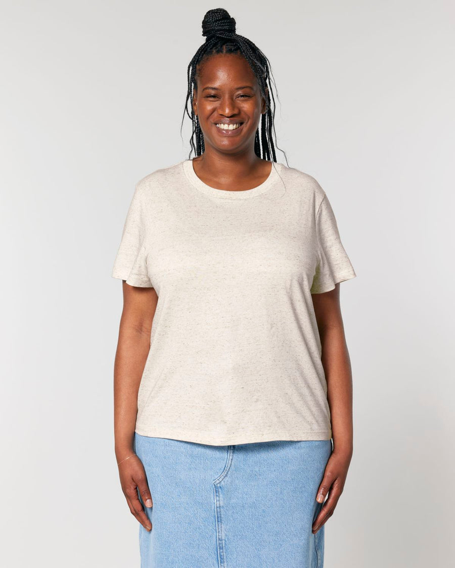 A person standing against a plain background, smiling, wearing a light-colored Stanley/Stella STTW172 Stella Muser The Iconic Womens T-Shirt and a blue denim skirt.