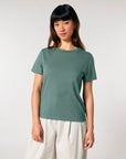 A person with long hair stands facing the camera, wearing a green Stanley/Stella STTW172 Stella Muser The Iconic Womens T-Shirt made of organic cotton and white pants, against a plain light background.
