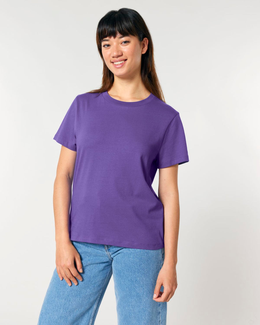 A person wearing a purple STTW172 Stella Muser The Iconic Womens T-Shirt by Stanley/Stella and jeans stands against a plain white background.