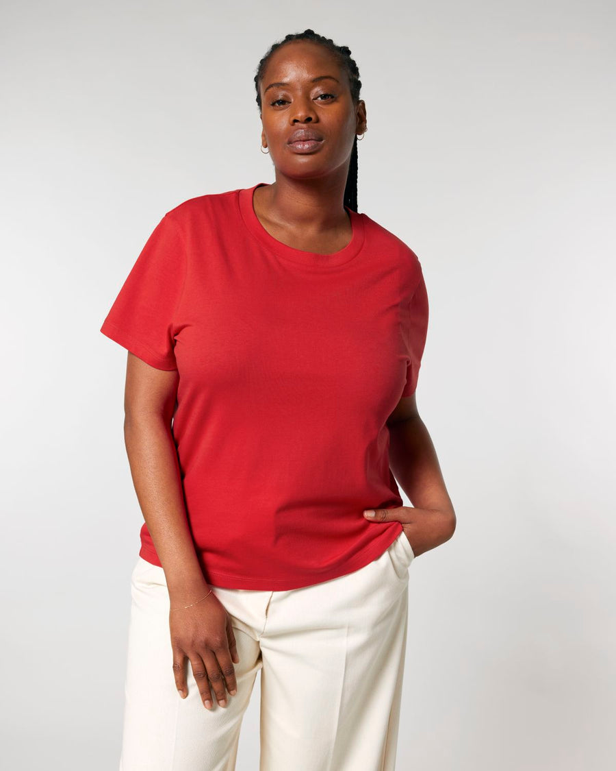 A woman wearing a red STTW172 Stella Muser The Iconic Women's T-Shirt by Stanley/Stella crafted from organic cotton and off-white pants stands against a plain background with one hand in her pocket.