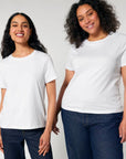 Two women wearing plain white STTW172 Stella Muser The Iconic Womens T-Shirts by Stanley/Stella and blue jeans stand side by side, smiling at the camera against a neutral background.