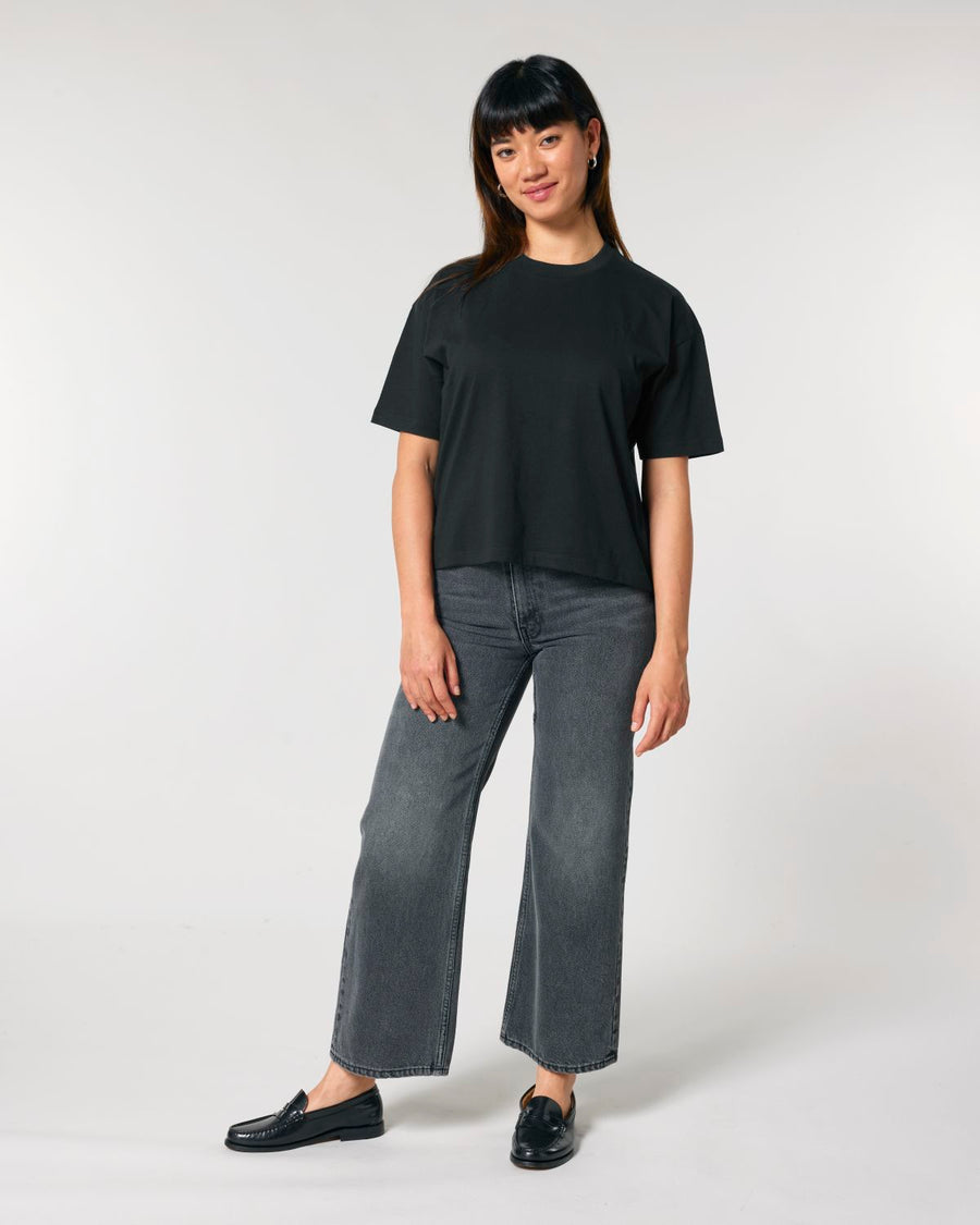 Woman standing in a studio posing in a casual black, STTW175 Stella Nova Womens Boxy T-Shirt made of organic cotton and gray jeans with black shoes.