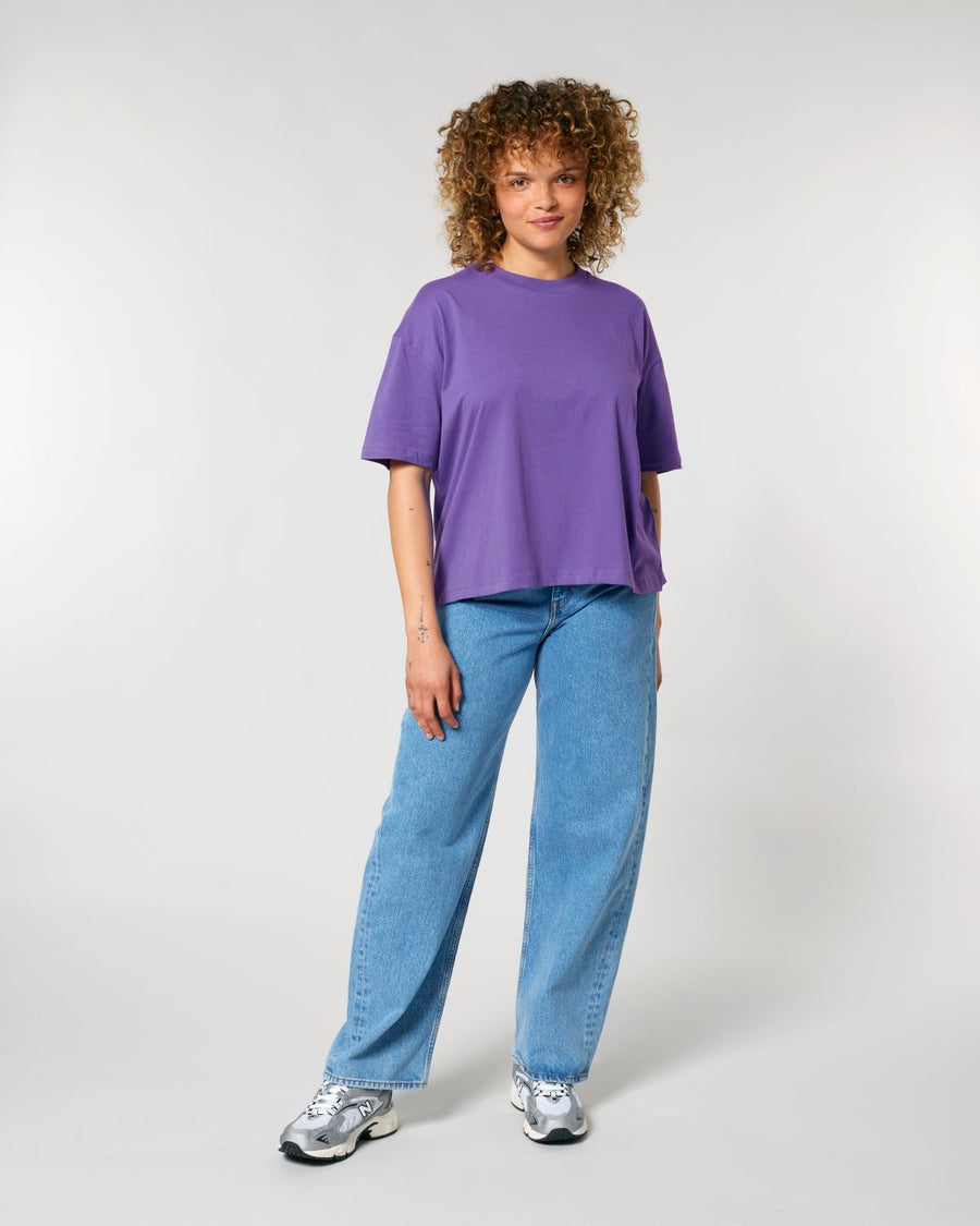 Woman standing in a relaxed pose wearing a STTW175 Stella Nova Women's Boxy T-Shirt by Stanley/Stella made of organic cotton and blue jeans with silver sneakers.